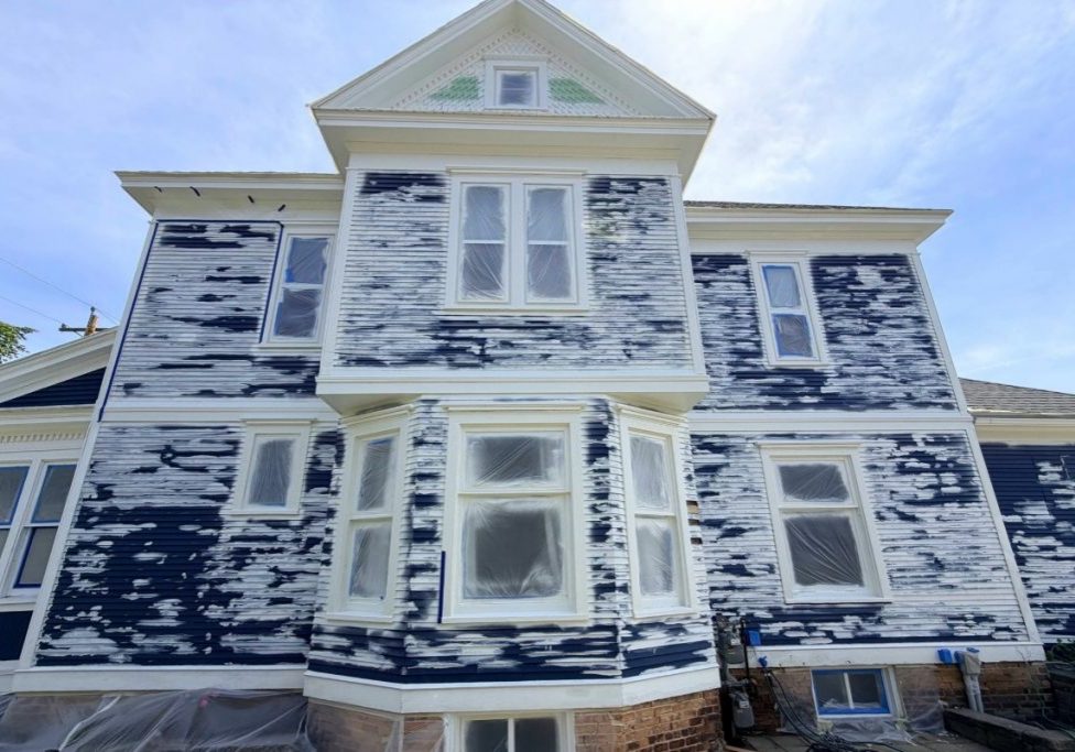 A large older two-story home with heavily peeling exterior paint scraped back to bare wood and primer during prep work, showing what happens when exterior paint reaches the end of how long it lasts.